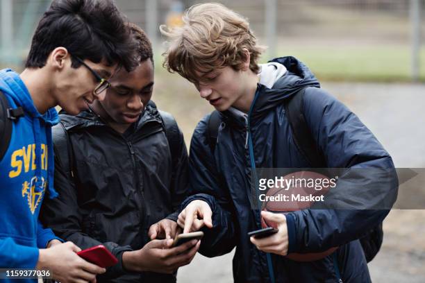 friends looking in teenage boy's phone while standing with ball on playing field - 3 teenagers mobile outdoors stock-fotos und bilder