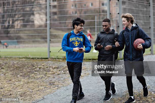 happy friends using mobile phones while walking on street against sports court - 3 teenagers mobile outdoors stock-fotos und bilder