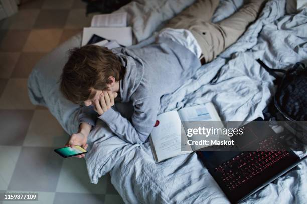 high angle view of teenage boy using mobile phone while lying by books and laptop on bed at home - boys bedroom stock pictures, royalty-free photos & images