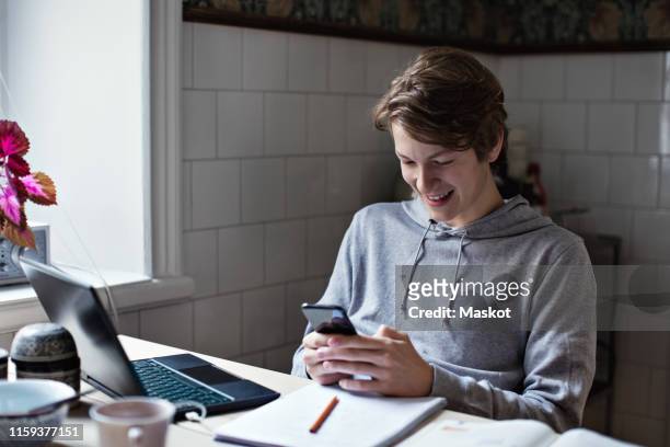 smiling social media addicted teenage boy using mobile phone while doing homework at table - addiction mobile and laptop stock-fotos und bilder