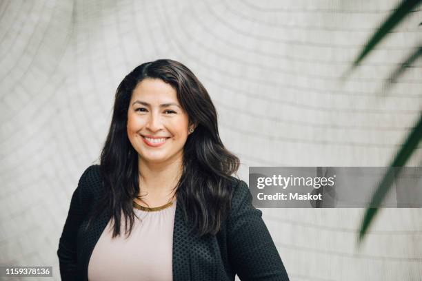 portrait of smiling businesswoman standing against wall at office - hispanic businesswoman stock pictures, royalty-free photos & images