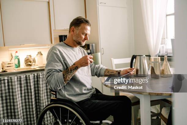 tattooed mature man reading mail while holding coffee cup on wheelchair at home - rolstoel stockfoto's en -beelden