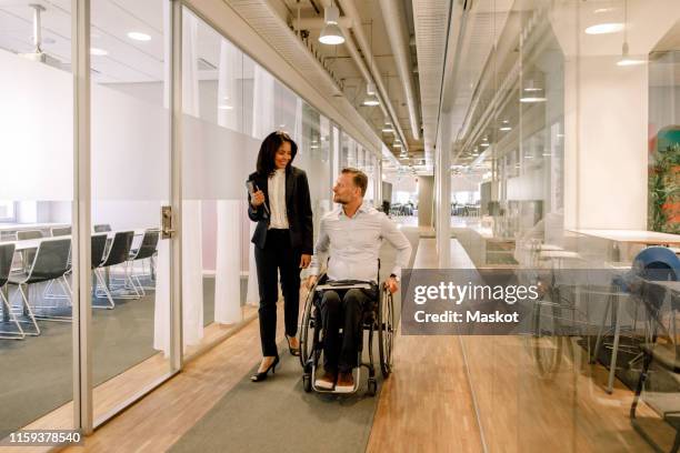 smiling businesswoman looking at disabled colleague sitting on wheelchair in corridor at work place - differing abilities female business stock-fotos und bilder