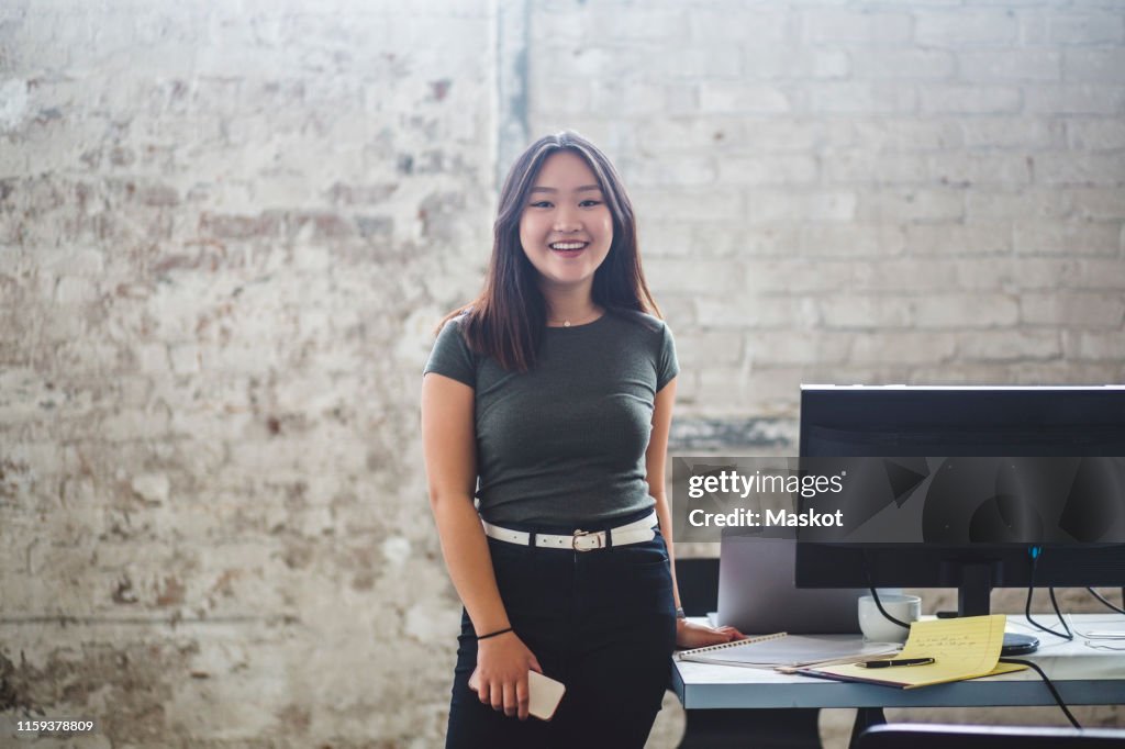 Portrait of smiling female computer programmer standing beside desk at creative office
