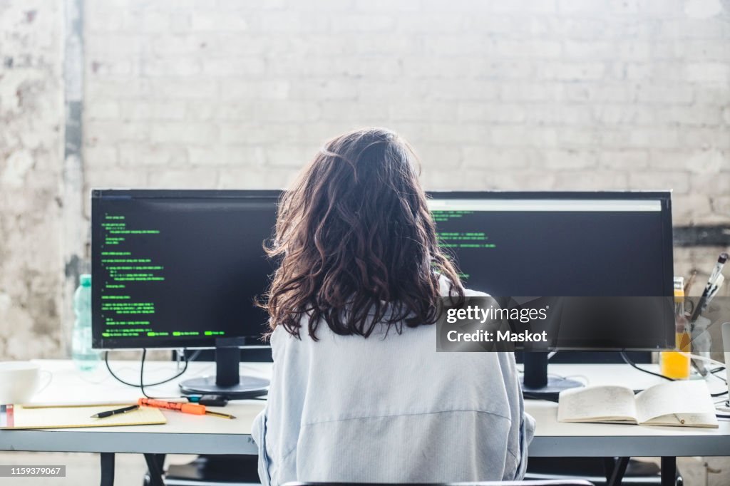 Rear view of female computer hacker coding at desk in creative office