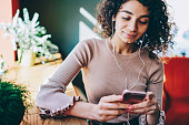 Attractive hipster girl listening audio book via mobile application and electronic headphones indoors, beautiful woman 20s updating notification on smartphone resting on leisure enjoying music