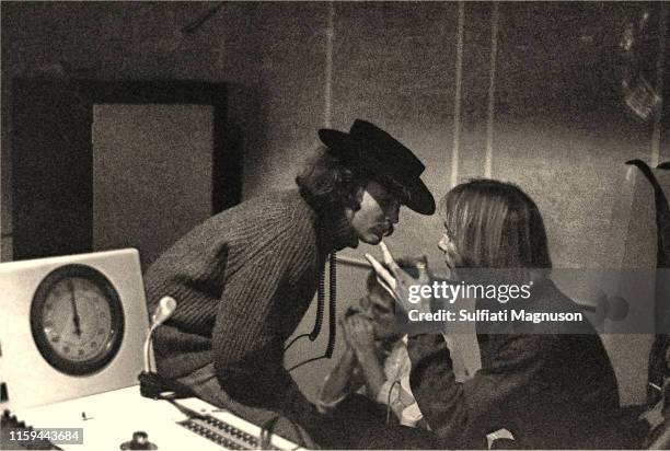 David Crosby talking to Stephen Stills while John Sebastian plays the harmonica, seen from the recording booth, looking into the control room.