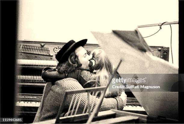 David Crosby kissing Joni Mitchell on the cheek while both are seated at an upright piano, seen from the back, through the control room window at the...
