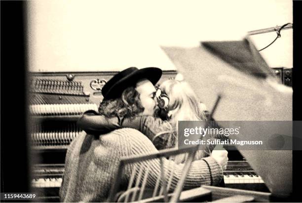 David Crosby holding Joni Mitchell while both are seated at an upright piano, seen from the back, through the control room window at the recording...