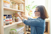 Interior of wooden pantry with products for cooking. Adult woman taking kitchenware and food from the shelves
