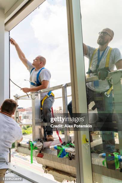 team of blue-collar workers replacing a broken window in the office building - cleaning the frame after the old broken glass has been removed. high-altitude work on the lifting platform which is placed outside. - replacement windows stock pictures, royalty-free photos & images