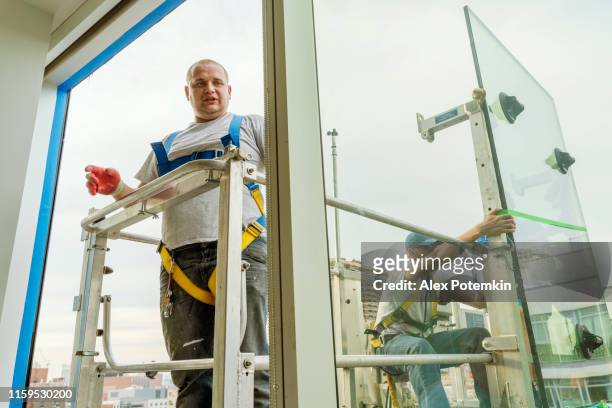 team of blue-collar workers replacing a broken window in the office building - applying a fresh sealant for a new glass installation. high-altitude work on the lifting platform which is placed outside. - replacement stock pictures, royalty-free photos & images