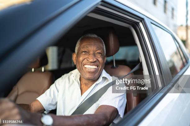 homme aîné de sourire conduisant une voiture et regardant l'appareil-photo - black car photos et images de collection