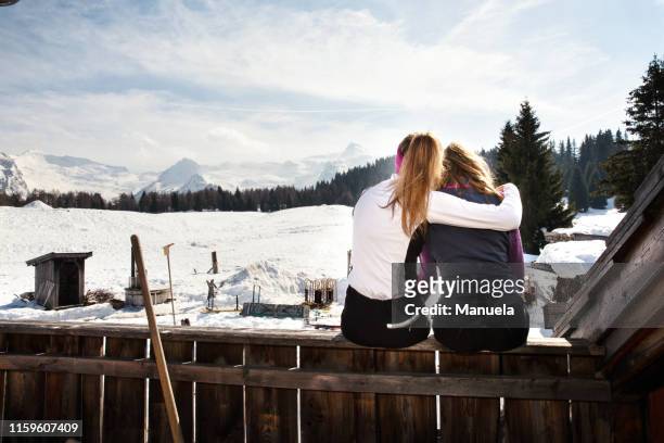 two teenage girl skiers sitting on cabin roof terrace in snow covered landscape, rear view, tyrol, styria, austria - styria stock-fotos und bilder