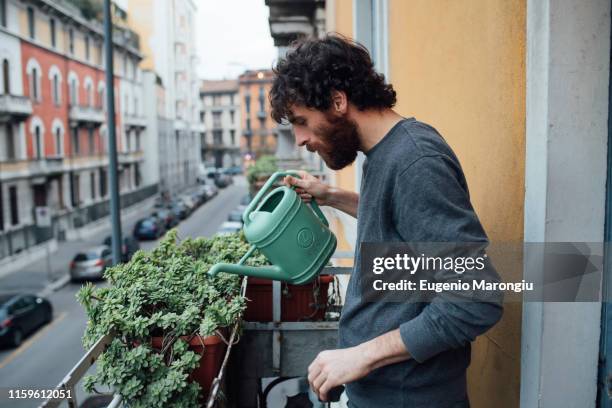 bearded young man watering plants on balcony - balkon pflanzen stock-fotos und bilder