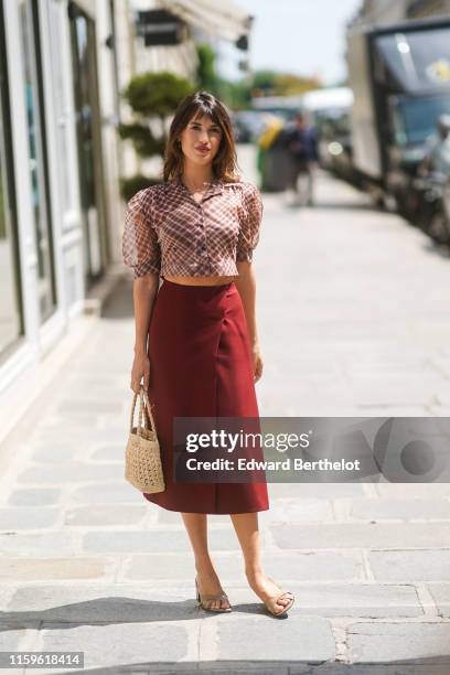 Jeanne Damas wears a Dior puff sleeves brown and red checkered crop shirt, a burgundy wrap-over skirt, a woven sisal bag, taupe-color mules , outside...