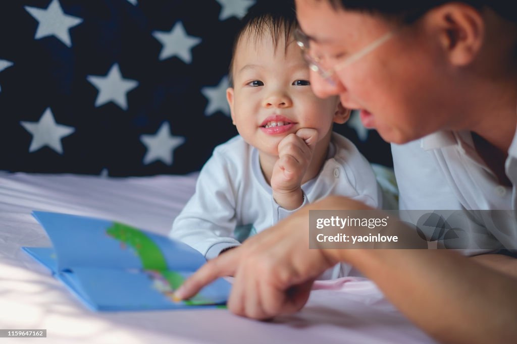 Father and Cute little Asian 2 - 3 years old toddler boy child reading bedtime story book, lying in the bed at home