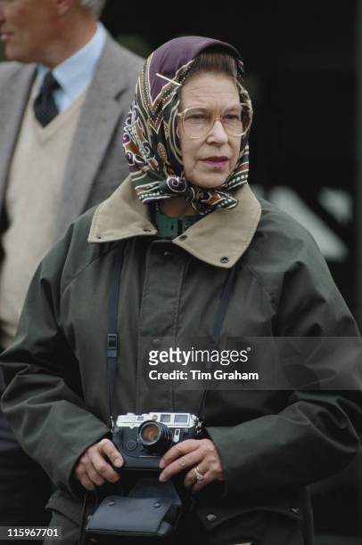 Queen Elizabeth II, wearing a headscarf and green waxed jacket, and holding a Leica camera, at the Royal Windsor Horse Show, held at Home Park in...