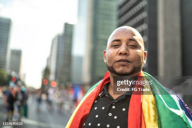 junge homosexuell mann blick auf die kamera und halten regenbogen-flagge während stolz parade - regenbogenfahne stock-fotos und bilder
