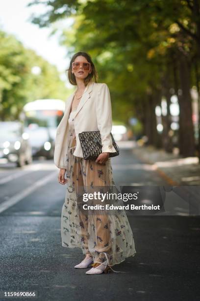 Guest wears sunglasses, earrings, a necklace, a white jacket, a Dior monogram bag, white transparent shoes, a fishnet embroidered ruffled long dress...