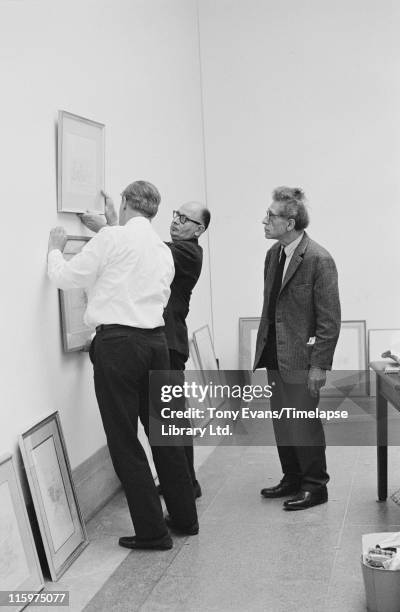 Swiss sculptor and painter Alberto Giacometti looks on as his friend Louis Clayeux and Robin Campbell hang a piece of work at the Tate Gallery,...