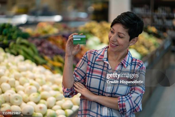 beautiful female customer looking at her loyalty card in a supermarket smiling - loyalty cards stock pictures, royalty-free photos & images