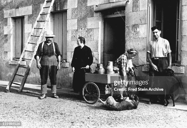 View of a farm family who pose beside a building with milk pail on a three-wheeled cart, France, 1935.