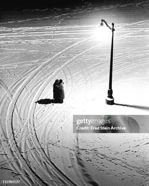 High angle, nighttime view of a couple as they stand under a street light and embrace on a snowy street corner, Paris, France, 1934.