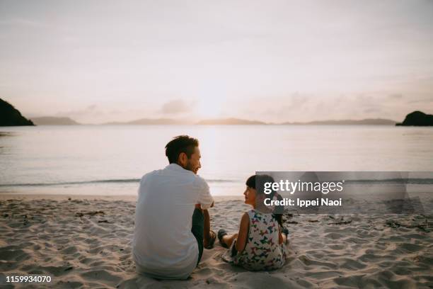 father and daughter sitting on beach and looking at each other at sunset - 2 people back asian imagens e fotografias de stock