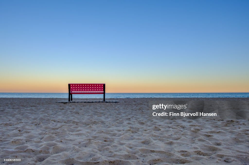Red empty bench at the beach at sunset, sand in foreground and ocean in backround