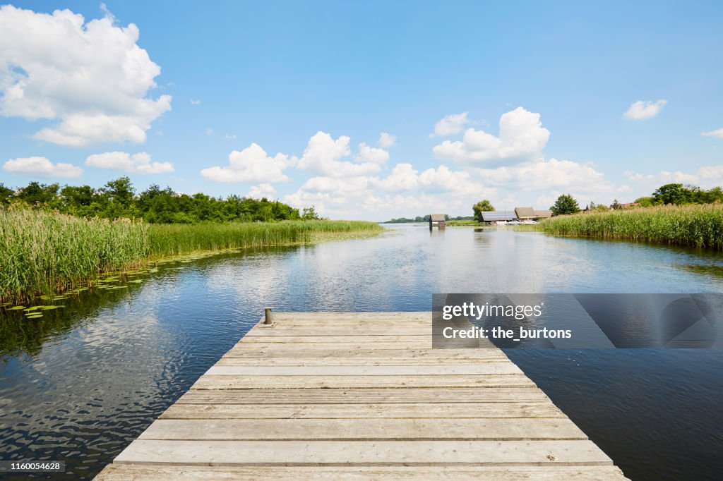 Jetty at idyllic lake with reed grass against blue sky, clouds are reflected in the water