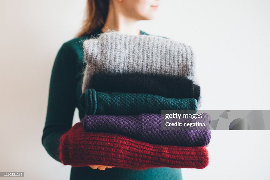 Young woman in green sweater holds stack of five different knitted warm winter sweaters