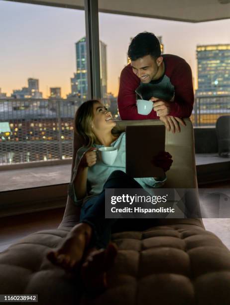 cute couple looking at something on tablet while talking and enjoying a cup of tea at home - hot latin nights stock pictures, royalty-free photos & images