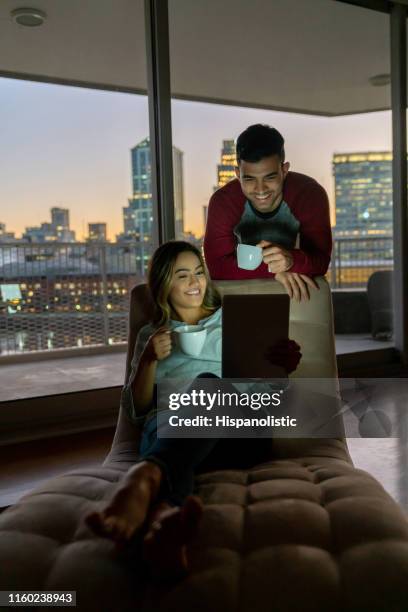 happy couple looking at something on tablet, woman relaxing on couch and man standing behind both enjoying a coffee smiling - hot latin nights stock pictures, royalty-free photos & images