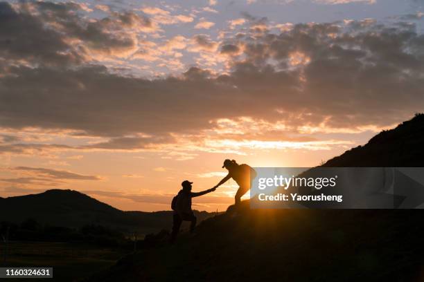 silhouette of people helping friend hike up a mountain in sunset,against cloudy sunset background - dan peak fotografías e imágenes de stock