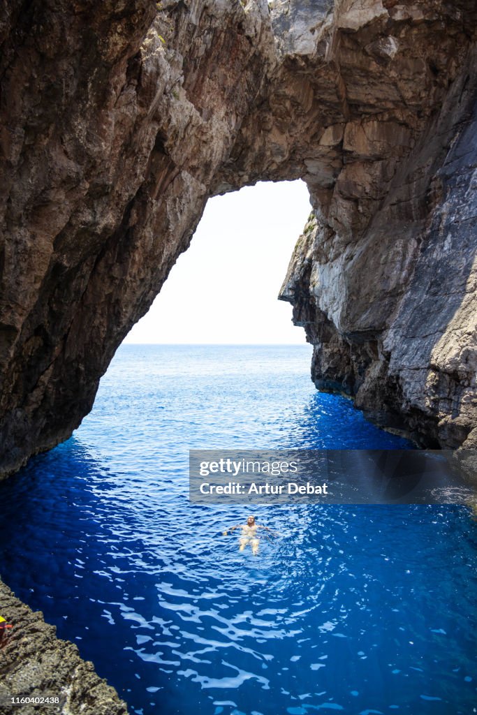 Traveler guy swimming with huge natural arch in Greece during summer.