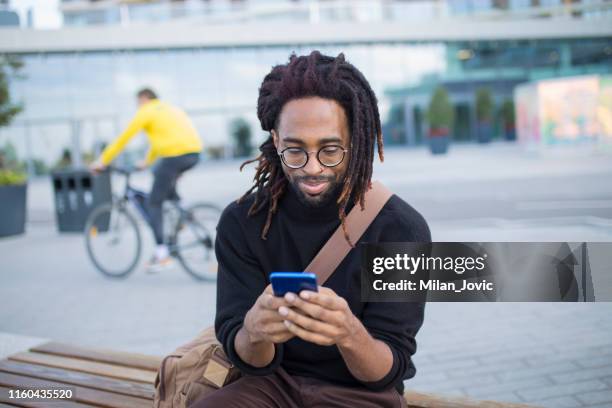 knappe man zittend op een bankje met een mobiele telefoon - dreadlocks stockfoto's en -beelden