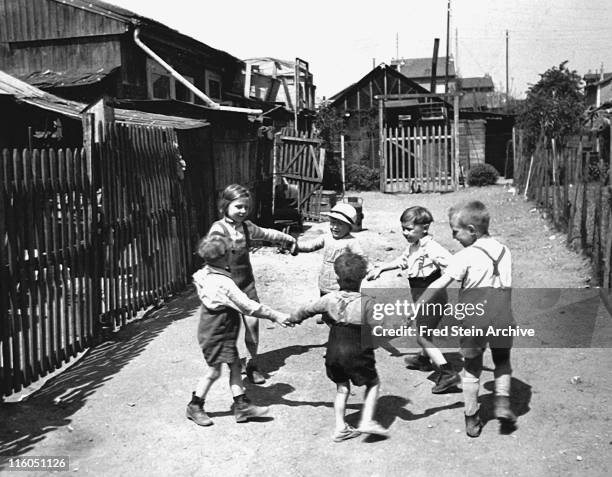 Group of children hold hands and play in a circle near the wooden fences of an alleyway, Paris, France, 1936.