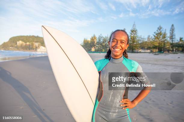 young indigenous australian girl on the beach in a wetsuit with her surfboard - australian aborigine culture stock pictures, royalty-free photos & images