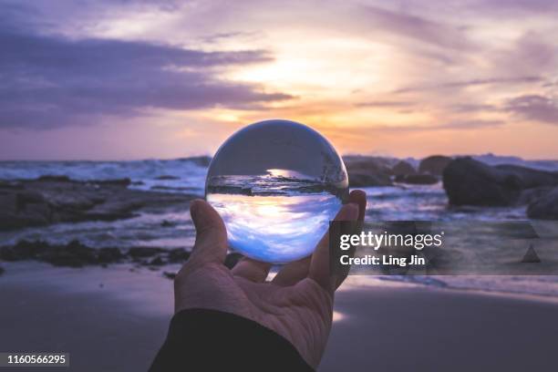 cropped hand of woman holding lens ball at beach against cloudy colorful sunset - glass sphere stock pictures, royalty-free photos & images