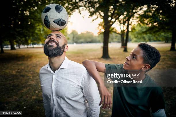 teenage boy looking at father balancing a soccer ball on his head in a park - man playing ball fotografías e imágenes de stock
