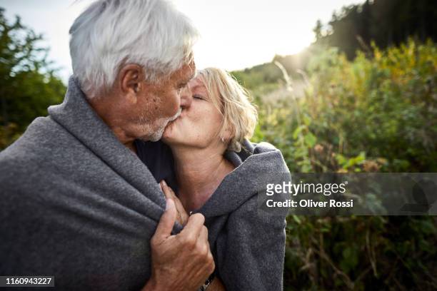 mature couple wrapped in a blanket kissing in the countryside - kuss stock-fotos und bilder