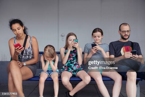 adults and children sitting on a bench distracted by mobile phones whilst younger child without a phone is completely fed up - last day bildbanksfoton och bilder