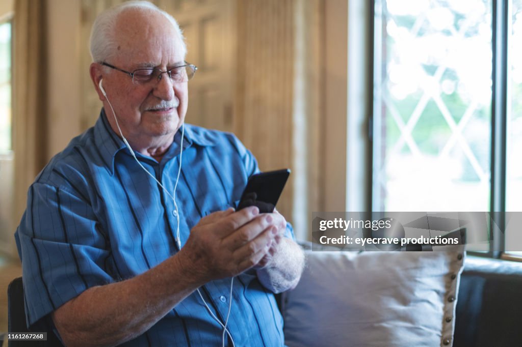 Mature retired adult male enjoying a life of leisure living inside on living room sofa listening to music and surfing the web on Smart phone technology