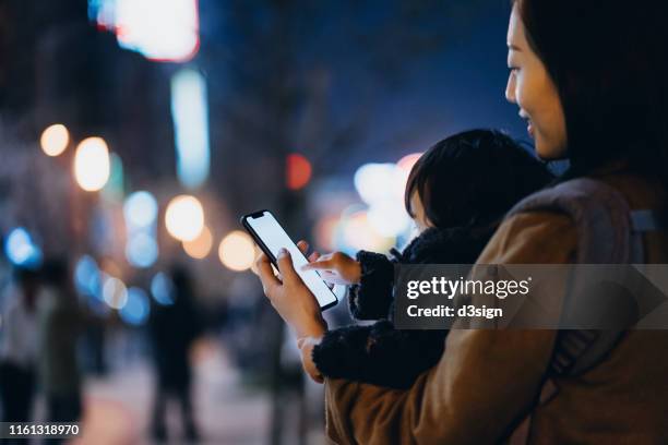 young asian mother carrying cute little daughter using smartphone in the city at night - mother and daughter on night street stockfoto's en -beelden