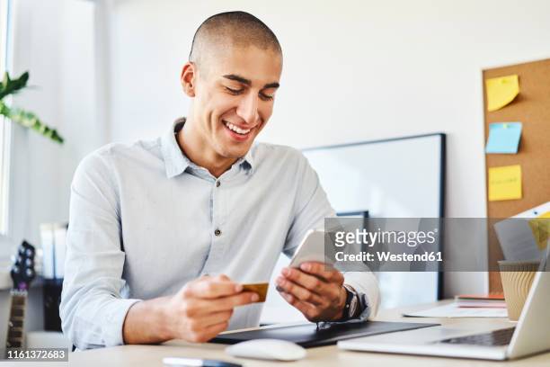 young man sitting in home office paying online with credit card and smartphone - business man sitting banking ストックフォトと画像