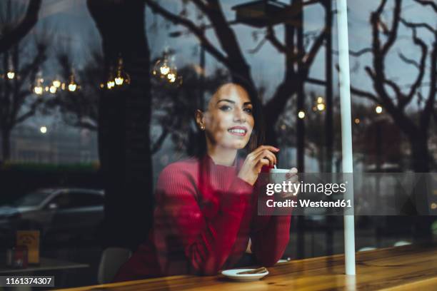 smiling young woman with cup of coffee behind windowpane in a cafe - café rouge photos et images de collection