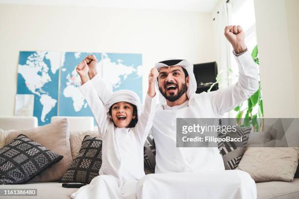 arab man looking tv at home during a sport event with his son - watching stock pictures, royalty-free photos & images