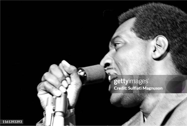 Otis Redding on stage, in a light colored, double breasted suit, mike in right hand, profile, spotlight above
