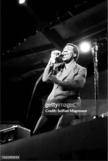 Otis Redding on stage, in a light colored, double breasted suit, mike in right hand, profile, spotlight above